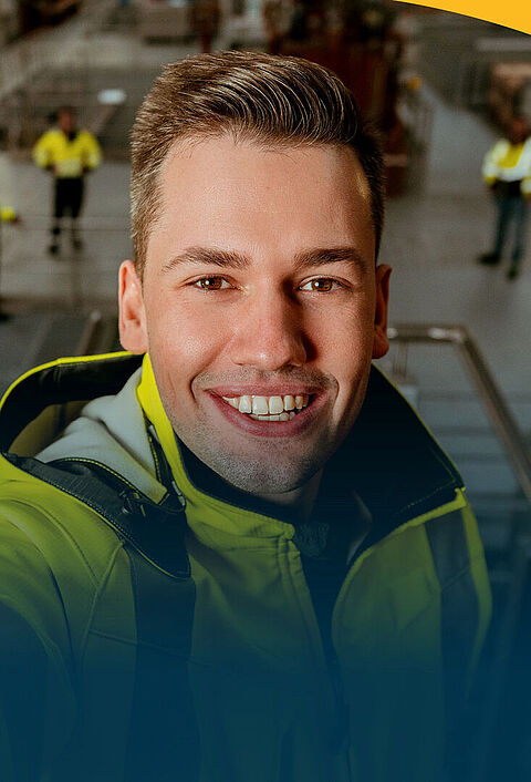 Ein Mann in Sicherheitsweste macht ein Selfie in einer Maschinenhalle. Im Hintergrund sieht man mehrere seiner Kollegen.