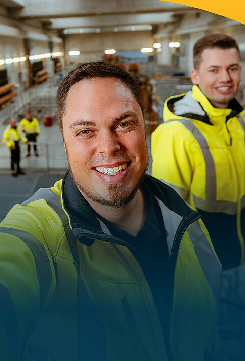 Ein lächelnder Mann macht ein Selfie in einer Maschinenhalle. Im Hintergrund sieht man mehrerer seiner Kollegen in Sicherheitswesten.