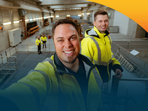 Ein lächelnder Mann macht ein Selfie in einer Maschinenhalle. Im Hintergrund sieht man mehrerer seiner Kollegen in Sicherheitswesten.