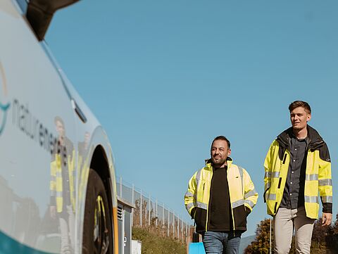 Zwei Männer in Arbeitskleidung laufen auf ein E-Auto von naturenergie zu