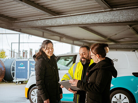 Mehrere Personen sprechen an einer E‑Auto-Ladestation neben einem geparkten Elektrofahrzeug