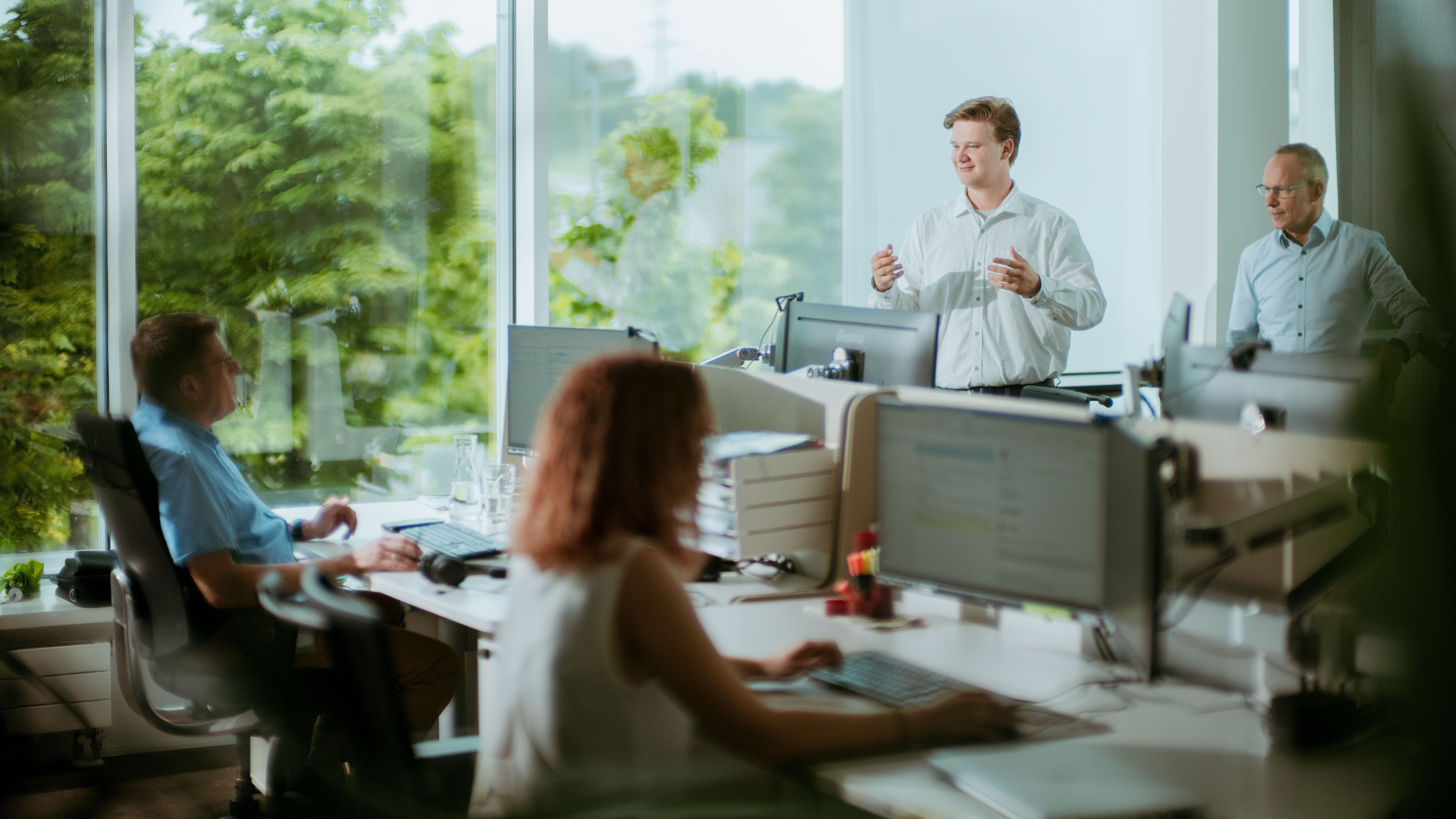 Ein Mitarbeiter präsentiert in einem hellen Großraumbüro etwas vor seinem Team, während Kolleginnen und Kollegen an ihren Arbeitsplätzen zuhören.