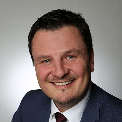 Porträt von Bürgermeister Adrian Schmidle im dunkelblauen Anzug mit weißem Hemd und bordeauxroter Krawatte vor neutralem Hintergrund.