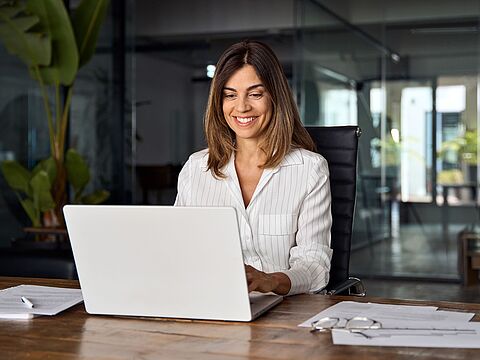 Person arbeitet an einem Laptop in einem modernen Büro mit Glaswänden und Pflanzen