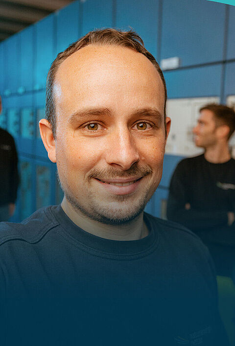 Ein Mann macht ein Selfie in einem technischen Anlagenraum mit Schaltschränken. Im Hintergrund sieht man zwei seiner Kollegen.