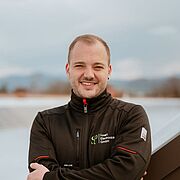Person mit schwarzer Jacke und blauen Jeans steht mit verschränkten Armen vor einer PV-Anlage und Landschaft mit Bergen und bewölktem Himmel