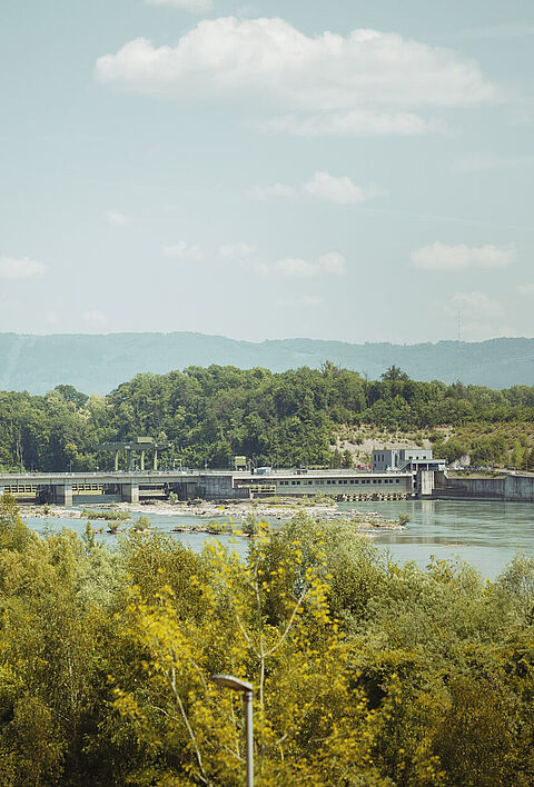 Wasserkraftanlage am Rhein, umgeben von Bäumen und einer hügeligen Landschaft.