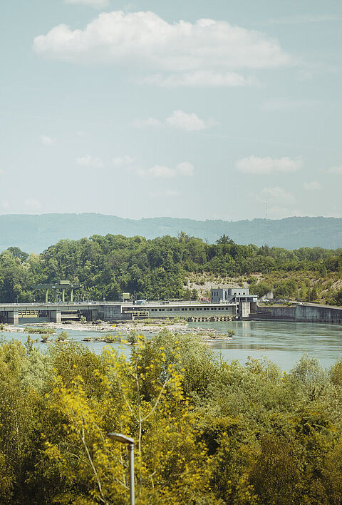 Wasserkraftanlage am Rhein, umgeben von Bäumen und einer hügeligen Landschaft.