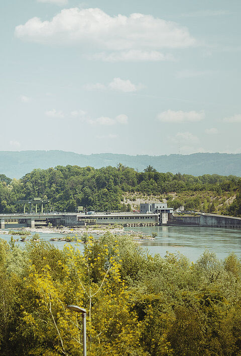 Wasserkraftanlage am Rhein, umgeben von Bäumen und einer hügeligen Landschaft.