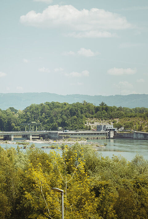 Wasserkraftanlage am Rhein, umgeben von Bäumen und einer hügeligen Landschaft.