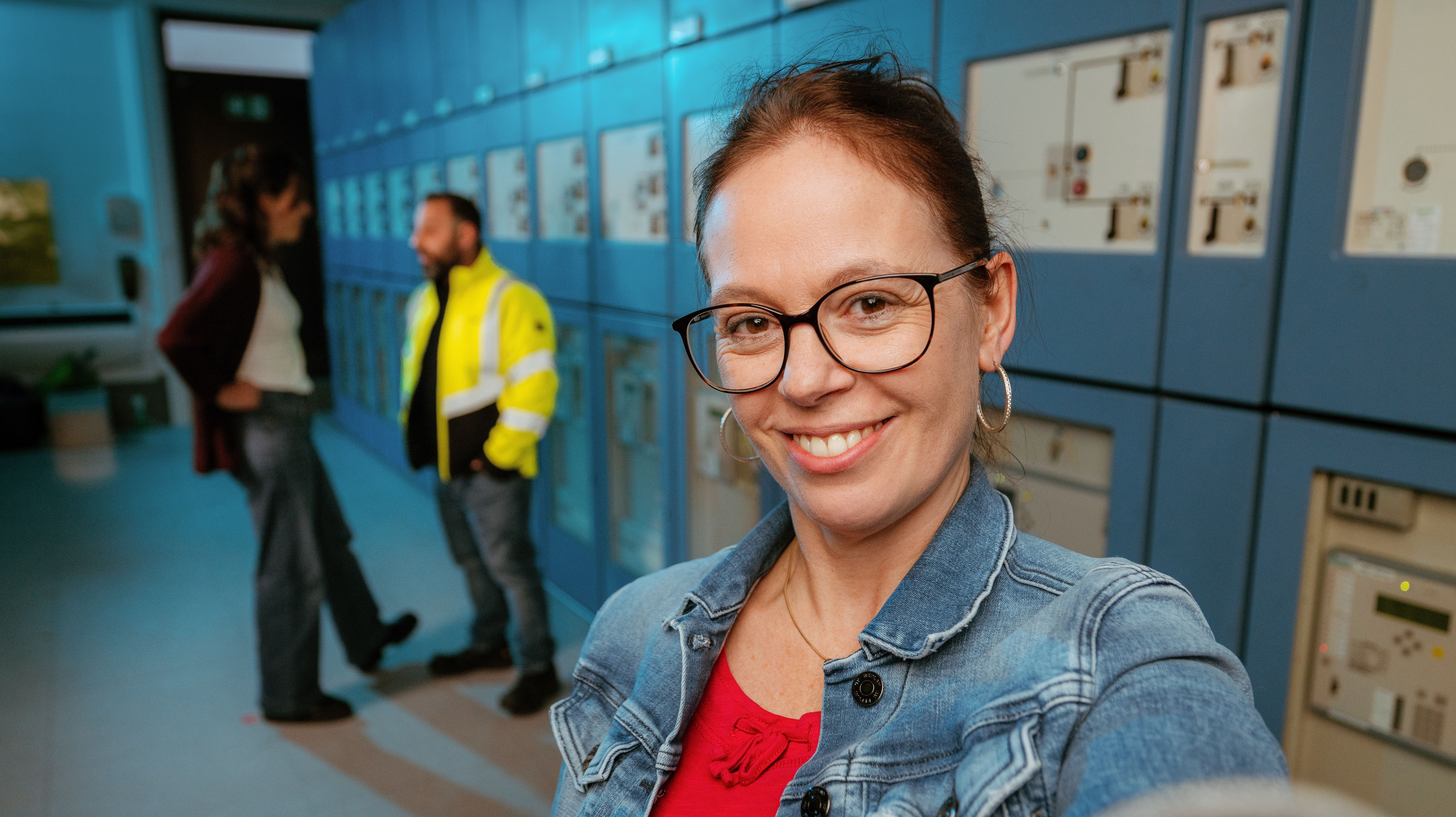 Eine Frau in Jeansjacke macht ein Selfie in einem technischen Anlagenraum mit Schaltschränken, während sich zwei Kollegen im Hintergrund unterhalten.