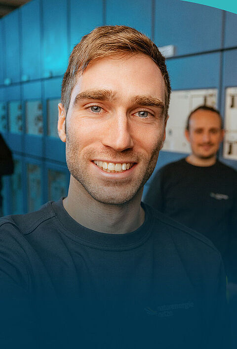 Ein lächelnder Mann macht ein Selfie in einem technischen Anlagenraum mit Schaltschränken. Zwei seiner Kollegen stehen im Hintergrund.
