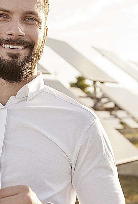 Glücklicher Geschäftsmann mit Brille in der Hand, der lächelt und die Kamera anschaut, während er vor Photovoltaik-Paneelen im Solarkraftwerk steht