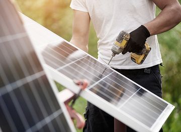 Techniker, der glänzende Solar-Photovoltaik-Module mit einem elektrischen Schraubendreher zusammenbaut, der im Freien auf hellgrünem, sonnigem, buntem, verschwommenem Bokeh-Hintergrund steht. Effizienz und Professionalität.
