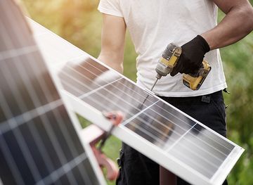 Wartung Solar-Photovoltaik-Module Techniker, der glänzende Solar-Photovoltaik-Module mit einem elektrischen Schraubendreher zusammenbaut, der im Freien auf hellgrünem, sonnigem, buntem, verschwommenem Bokeh-Hintergrund steht. Effizienz und Professionalität.