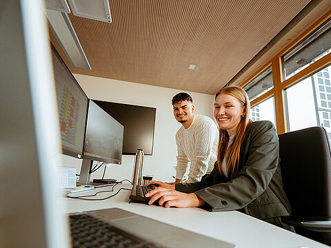 Zwei Personen arbeiten gemeinsam an einem Schreibtisch mit Monitor und Laptop.