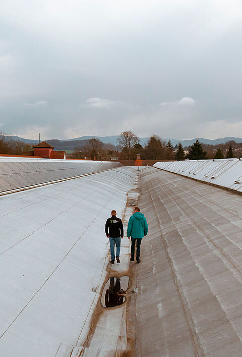 Zwei Personen auf einem großen Dach mit Solarpaneelen, im Hintergrund Bäume und Gebäude unter bewölktem Himmel.