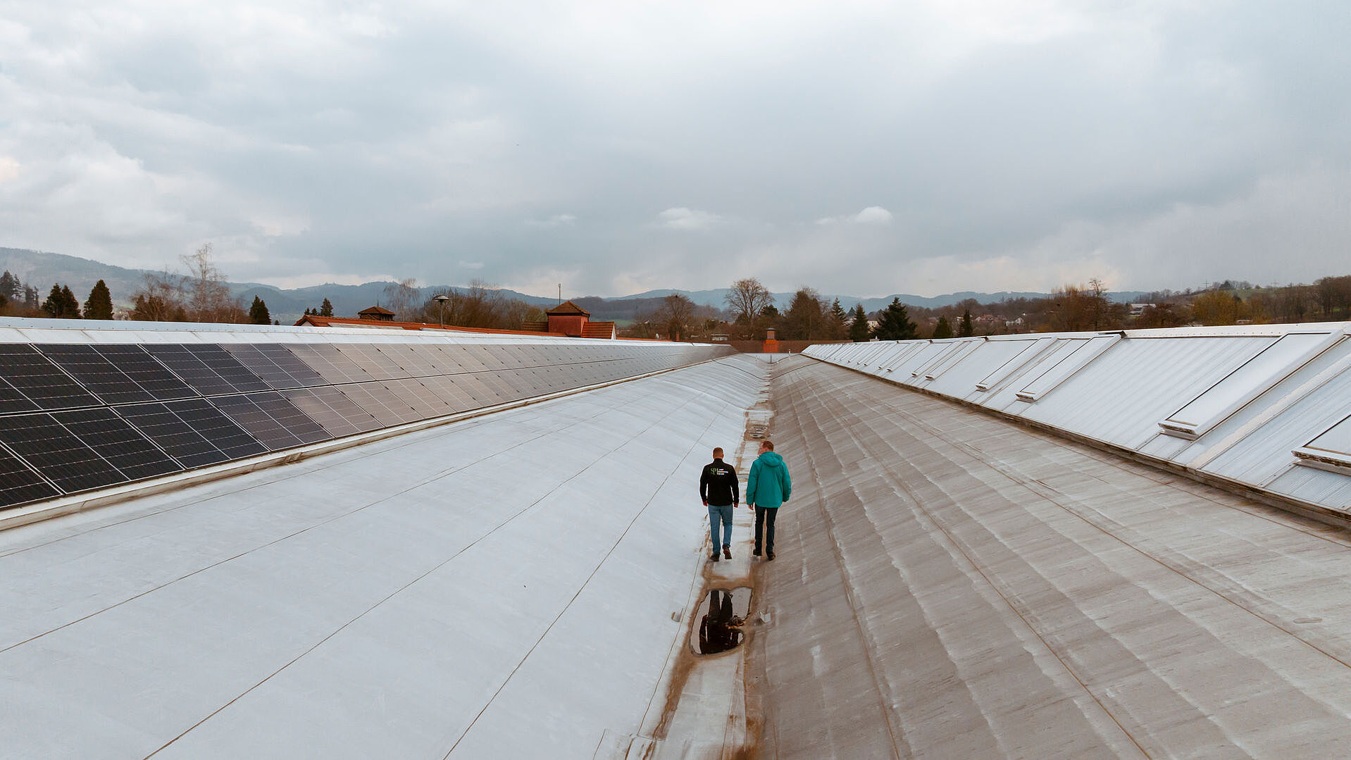 Zwei Personen auf einem großen Dach mit Solarpaneelen, im Hintergrund Bäume und Gebäude unter bewölktem Himmel.
