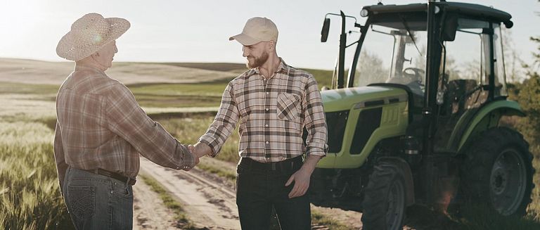 Der Mann nähert sich dem alten Bauer und schüttelt die Hände. Sie lächeln. Hinter ihnen steht ein Traktor. Die Sonne scheint hell.