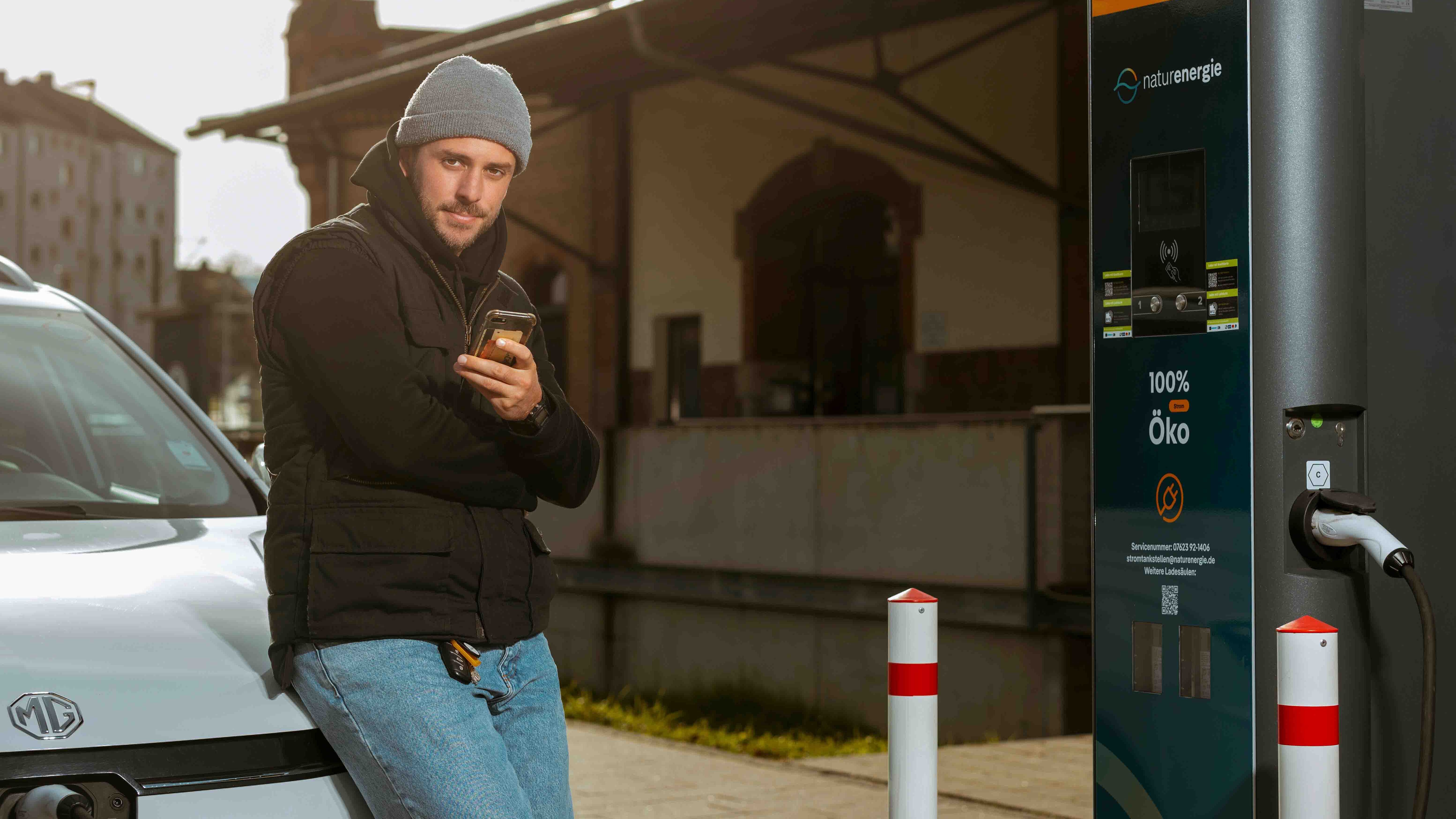 Person lehnt am Elektroauto und schaut auf Smartphone, Fahrzeug wird geladen