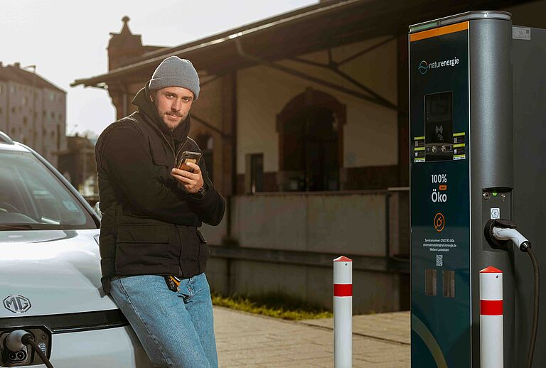 Person lehnt am Elektroauto und schaut auf Smartphone, Fahrzeug wird geladen