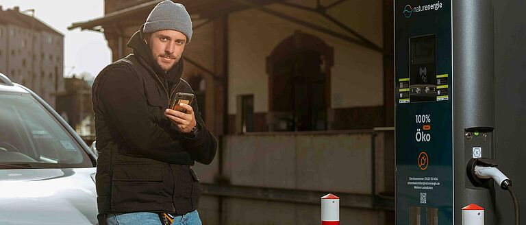 Person lehnt am Elektroauto und schaut auf Smartphone, Fahrzeug wird geladen