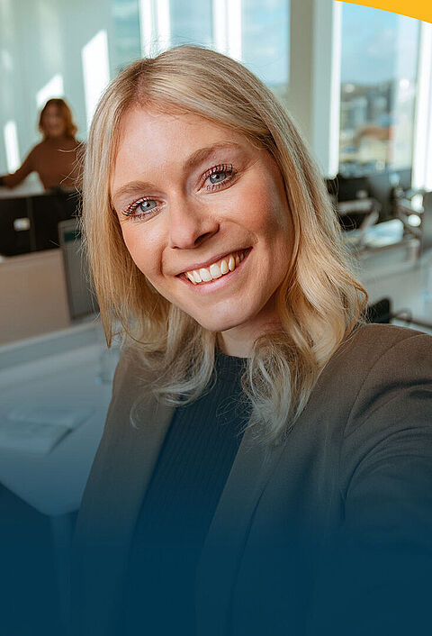 Eine lächelnde Frau macht ein Selfie im Büro. Im Hintergrund sieht man vier ihrer Kolleginnen und Kollegen.