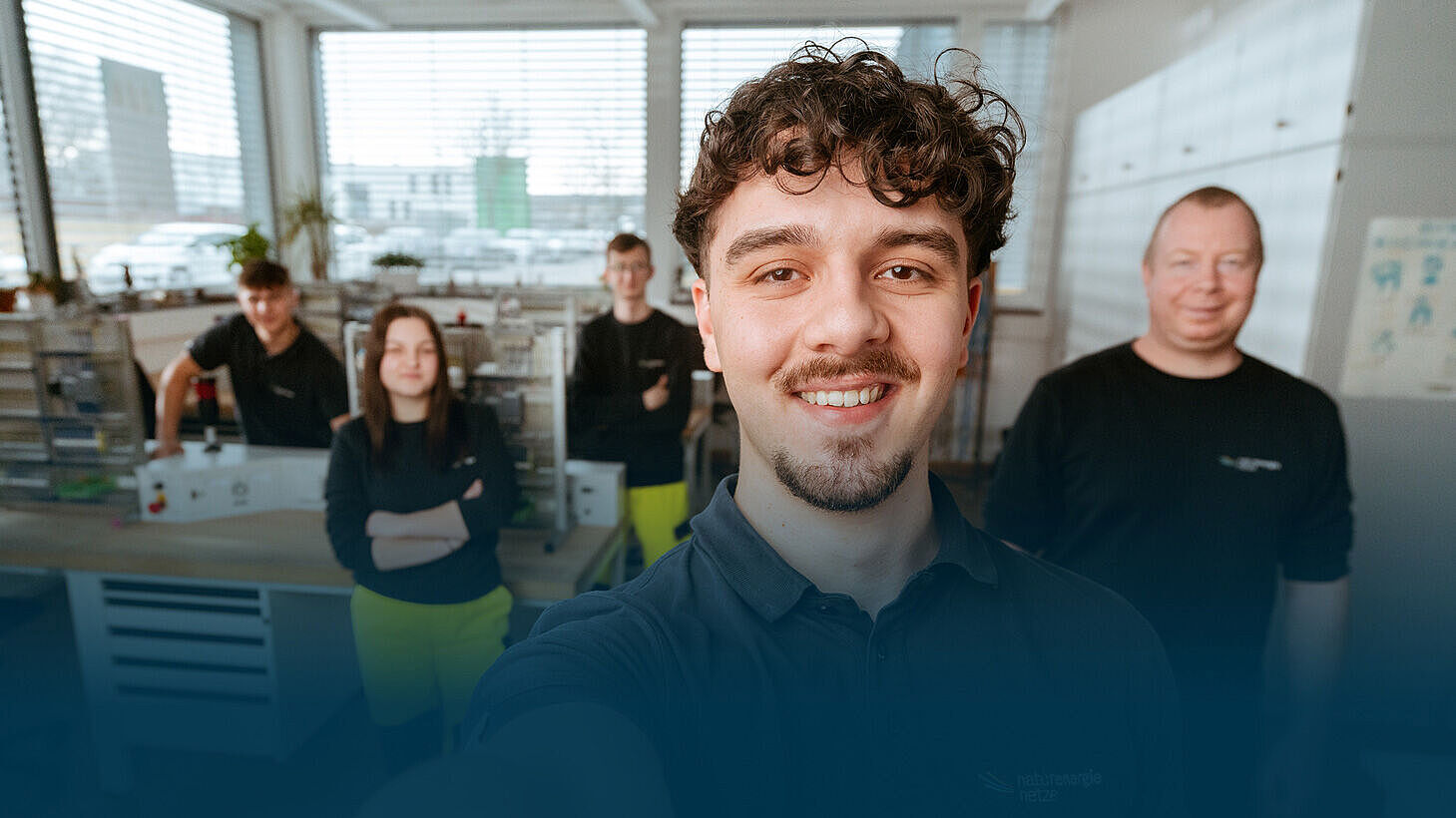 Ein Junge macht ein Selfie in einem Büro. Drei weitere Jugendliche und ein älterer Mitarbeiter sind im Hintergrund zu sehen und blicken in die Kamera.