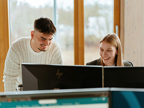 Zwei Personen arbeiten gemeinsam an einem Büroarbeitsplatz mit mehreren Bildschirmen.