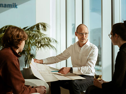 Drei Personen im Büro, eine reicht ein Dokument über den Tisch