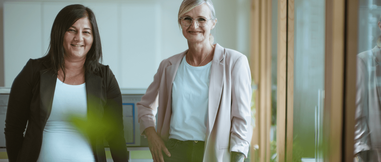 das Bild zeigt zwei Frauen ab der Hüfte, in einem Büro stehend