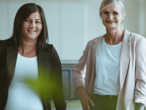 das Bild zeigt zwei Frauen ab der Hüfte, in einem Büro stehend