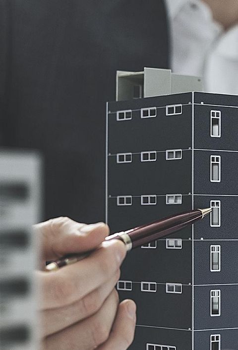 Ein Geschäftsmann im Anzug zeigt mit einem Kugelschreiber auf ein Modell eines Mehrfamilienhauses
