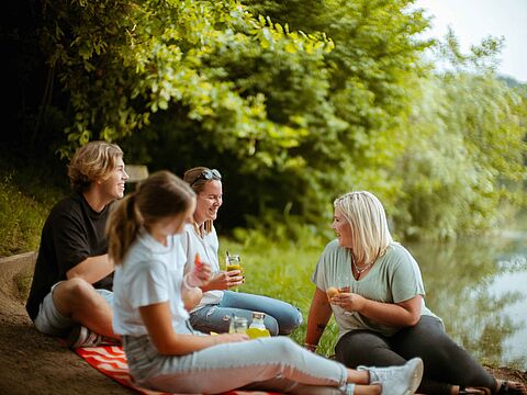 Vier Personen beim Picknick am Wasser in grüner Umgebung
