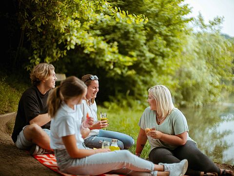 Vier Personen beim Picknick am Wasser in grüner Umgebung