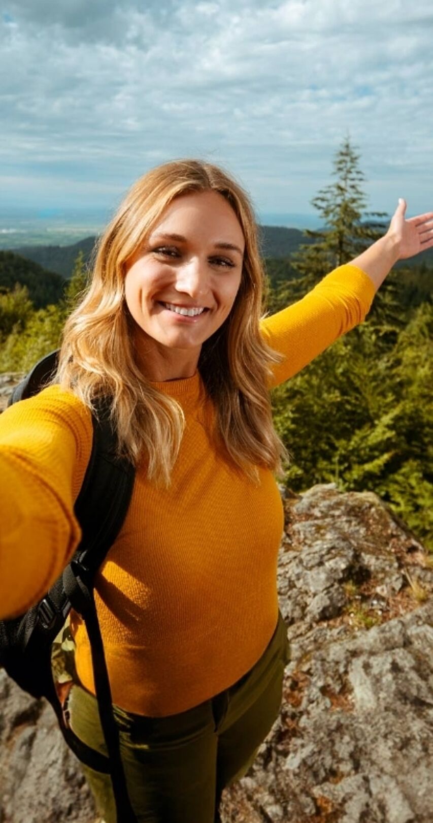 Person mit Rucksack steht mit ausgebreiteten Armen auf Felsvorsprung im Wald