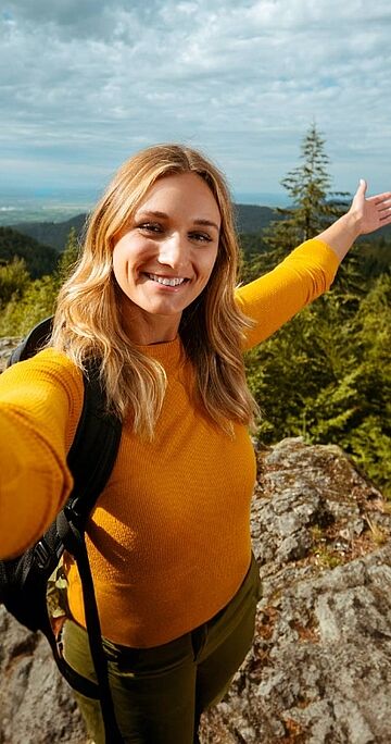Person mit Rucksack steht mit ausgebreiteten Armen auf Felsvorsprung im Wald