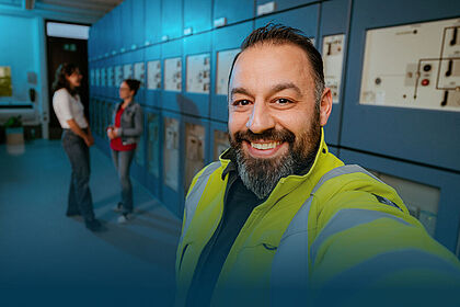 Ein lächelnder Techniker in einer gelben Warnjacke macht ein Selfie vor einer Reihe von elektrischen Schalttafeln in einem Technikraum. Im Hintergrund unterhalten sich zwei Kolleginnen.