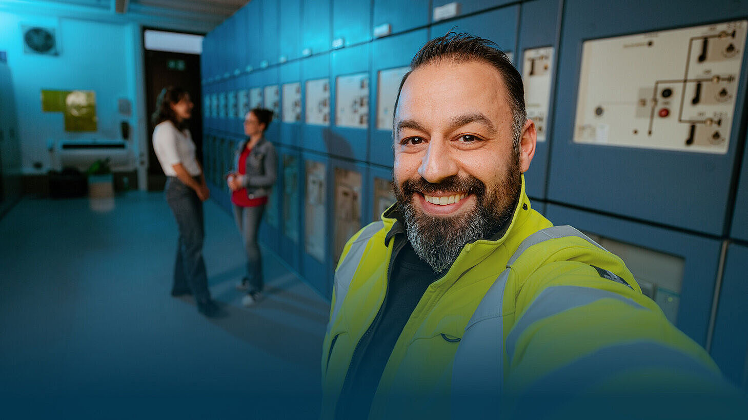 Ein lächelnder Techniker in einer gelben Warnjacke macht ein Selfie vor einer Reihe von elektrischen Schalttafeln in einem Technikraum. Im Hintergrund unterhalten sich zwei Kolleginnen.
