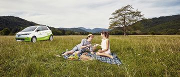 Paar Picknick my-e-car Ein Paar macht ein Picknick auf einer saftig grünen Wiese mit schöner Landschaft, im Hintergrund steht ein Elektrofahrzeug von my-e-car.