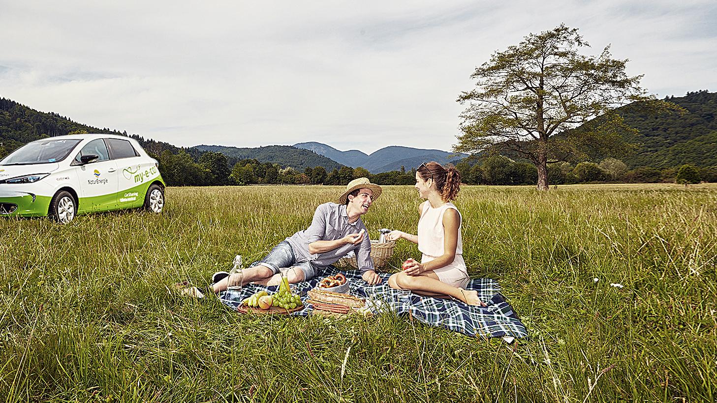 Mann und Frau picknicken auf der Wiese, im Hintergrund steht ein E-Auto