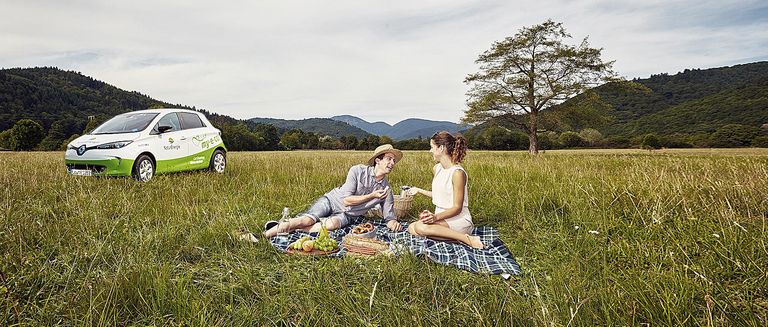 Ein Paar macht ein Picknick auf einer saftig grünen Wiese mit schöner Landschaft, im Hintergrund steht ein Elektrofahrzeug von my-e-car.