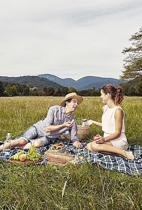 Ein Paar macht ein Picknick auf einer saftig grünen Wiese mit schöner Landschaft, im Hintergrund steht ein Elektrofahrzeug von my-e-car.