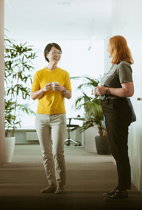 Das Bild zeigt drei Personen, die sich unterhalten. Die Personen stehen in einem Büro im Hintergrund sind Schreibtische und Pflanzen zu sehen. 