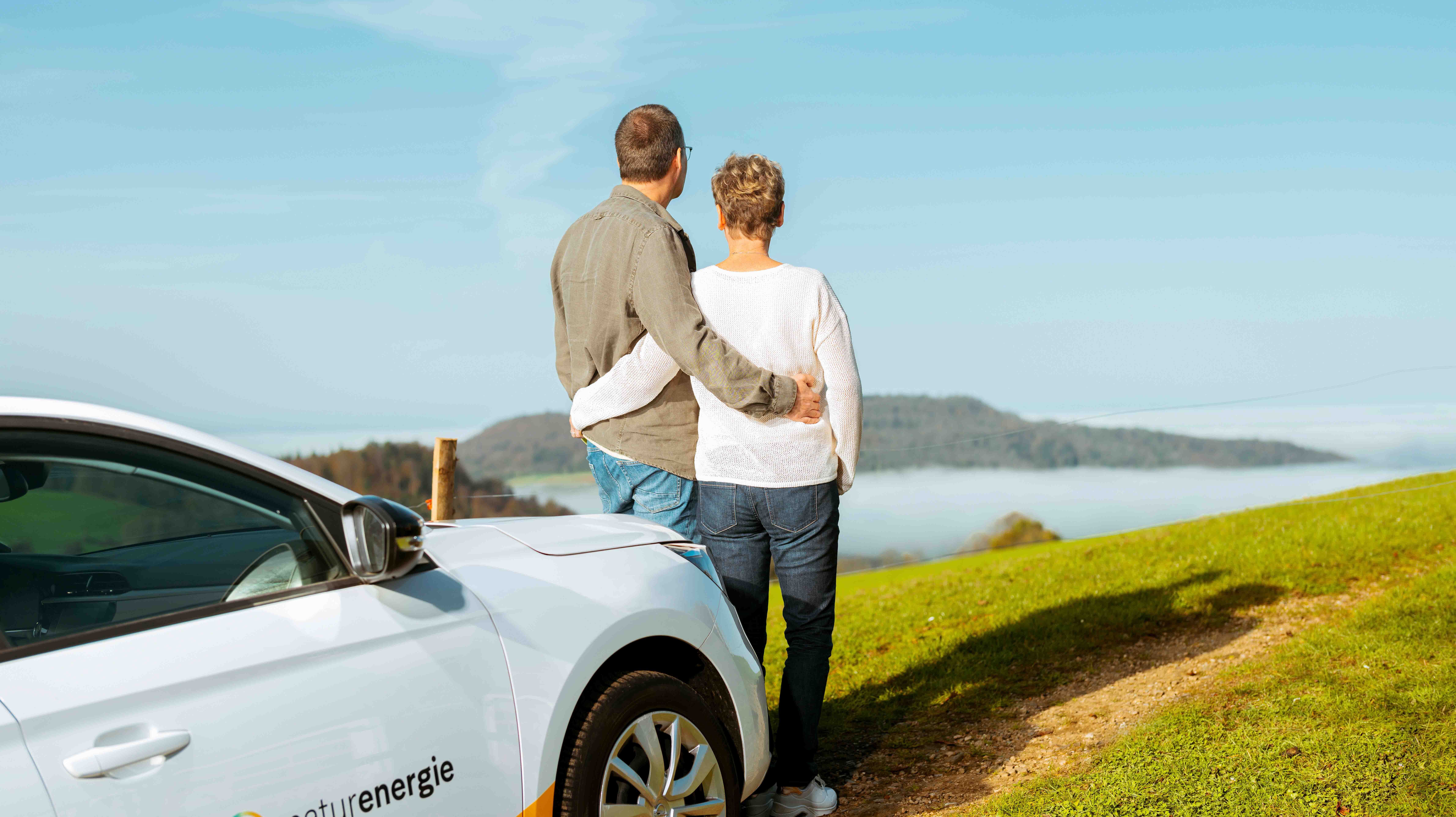 Zwei Personen stehen auf Hügel mit naturenergie-Auto, Blick auf Landschaft 