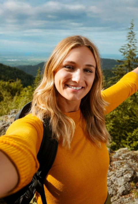 Person mit Rucksack macht Selfie auf Felsvorsprung in bewaldeter Landschaft