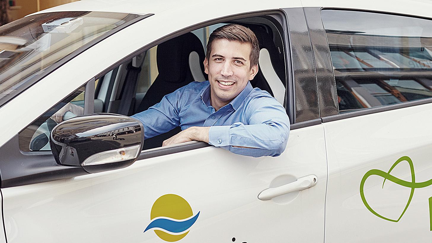 Ein Mann mit blauem Hemd lehnt aus dem Fahrerfenster und lacht in die Kamera. Er fährt ein Elektrofahrzeug von my-e-car. 