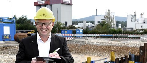 Leiter des Bereichs Wärme-und Energielösungen steht mit Schreibbrett und Bauhelm mitten auf einer Nahwärme-Baustelle mit Rohrleitungen vor dem Industriegebäude Evonik