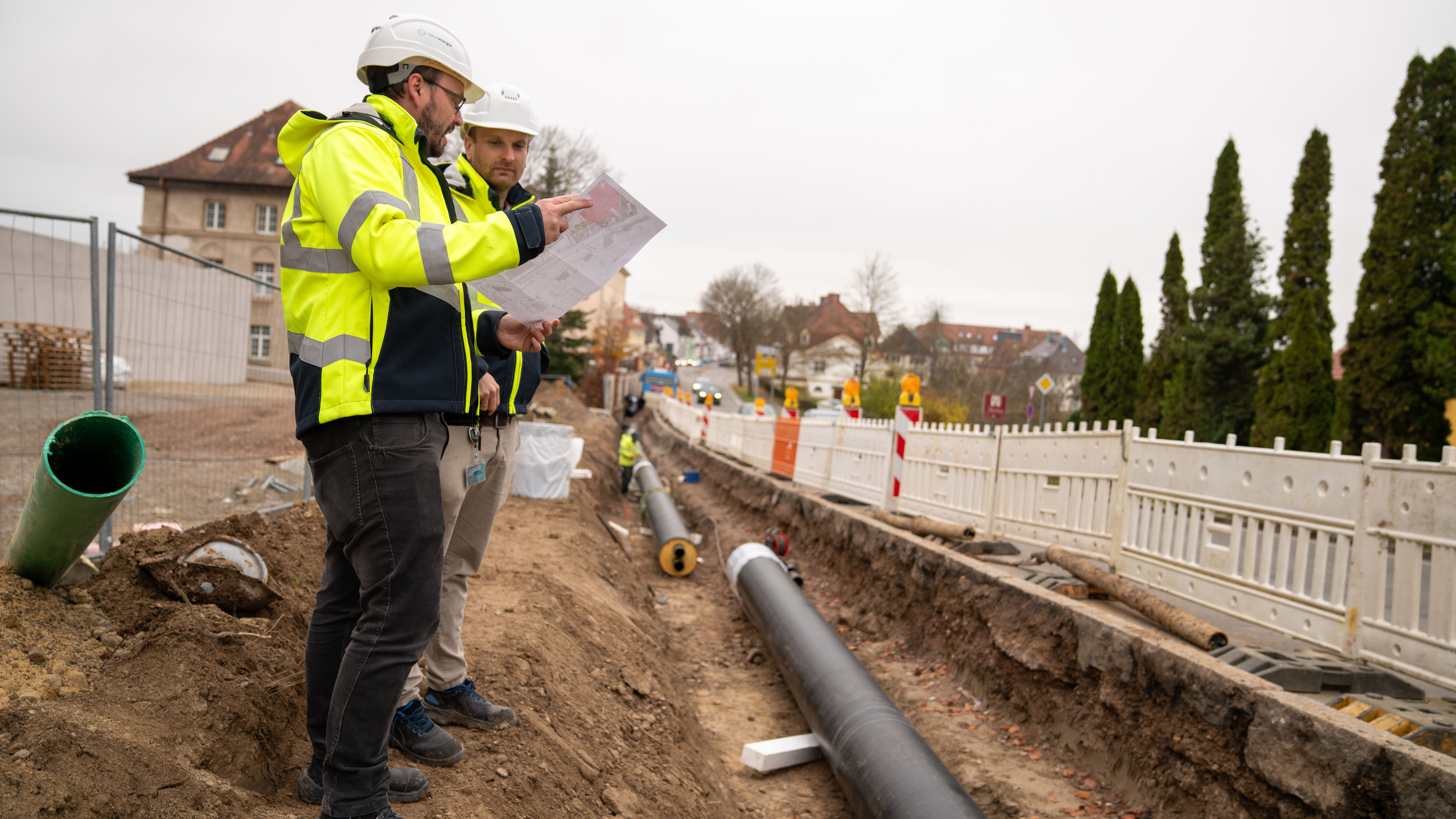 Zwei Bauingenieure mit Helmen besprechen Baupläne an einer Baustelle mit Rohrleitung im Graben.