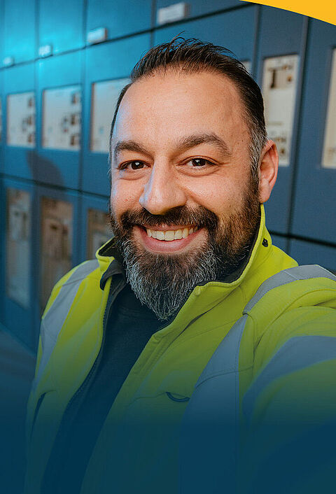 Ein lächelnder Techniker in einer gelben Warnjacke macht ein Selfie vor einer Reihe von elektrischen Schalttafeln in einem Technikraum. Im Hintergrund unterhalten sich zwei Kolleginnen.